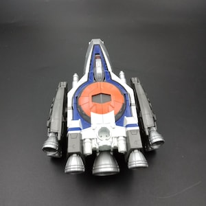 May include: A toy spaceship in silver, white, blue, and orange. The toy has multiple thrusters and a central circular design. The spaceship is set against a dark background.