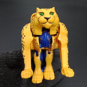 May include: A yellow and blue toy robot in the shape of a cheetah. The toy has black spots, green eyes, and is posed in a standing position. The toy is made of plastic and has articulated joints.