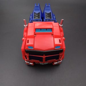 May include: A red and blue toy truck, viewed from above, with a rectangular body and two blue structures on top. The truck has a detailed front with black and blue accents and two silver exhaust pipes.