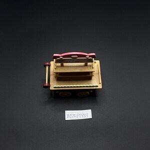 May include: A miniature wooden cart with a pink sign and two shelves. The cart has four wheels and a handle. The sign has text on it. The cart is made of light brown wood and has a dark brown trim. The background is black.