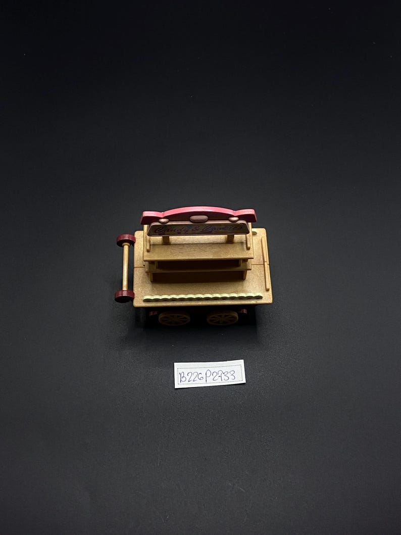 May include: A miniature wooden cart with a pink sign and two shelves. The cart has four wheels and a handle. The sign has text on it. The cart is made of light brown wood and has a dark brown trim. The background is black.