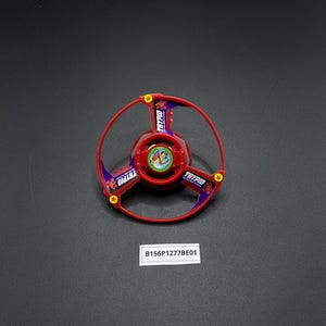 May include: Red and purple plastic toy top with a scorpion design and the word "TRYPIO" printed on it. The top is on a black surface.
