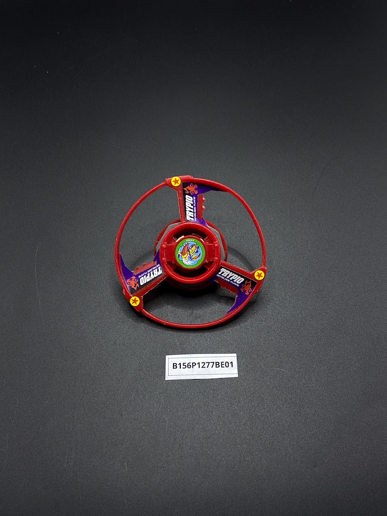 May include: Red and purple plastic toy top with a scorpion design and the word "TRYPIO" printed on it. The top is on a black surface.