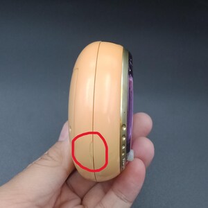 May include: A peach-colored, handheld electronic device with a gold-colored trim. The device has a purple screen and several small buttons. The device is held in a hand.
