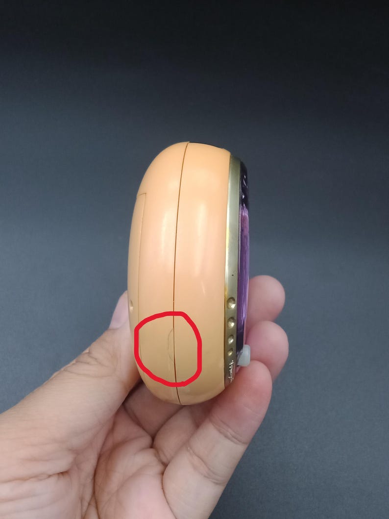 May include: A peach-colored, handheld electronic device with a gold-colored trim. The device has a purple screen and several small buttons. The device is held in a hand.