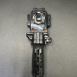 May include: A black toy launcher with a textured handle and a circular opening at the top. The launcher has several orange accents and a small loop on the side. The handle has a series of vertical slots.