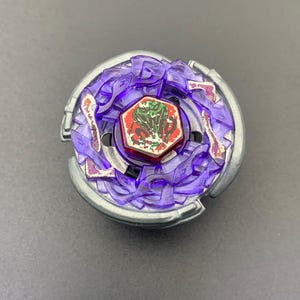 Puede incluir: Un juguete de peonza circular con una base de metal plateado y un anillo de plástico morado translúcido con detalles intrincados y dentados. En el centro, un emblema hexagonal rojo muestra un gráfico de una cobra verde con desgaste visible.