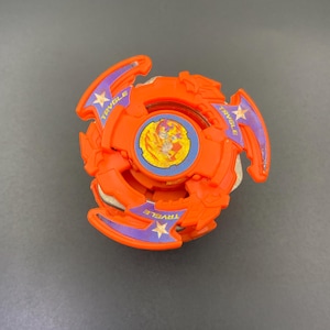 May include: An orange plastic spinning top toy featuring three protruding arms with purple accents, gold star graphics, and the text TRYGLE printed on each arm. The center of the toy displays a circular emblem with a yellow and red graphic design.