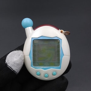 Puede incluir: Un dispositivo digital portátil blanco y azul para mascotas con una pequeña pantalla y tres botones turquesa. El dispositivo tiene una parte superior roja y blanca y una esfera azul en la parte superior. Un anillo dorado está unido al lateral.