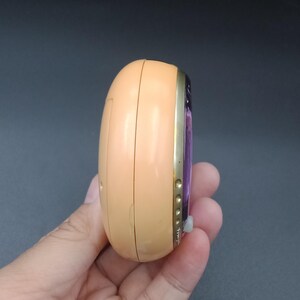May include: A peach-colored, oval-shaped electronic device with a gold-colored trim. The device has a small, white button and a purple screen. The device is held in a hand against a dark background.