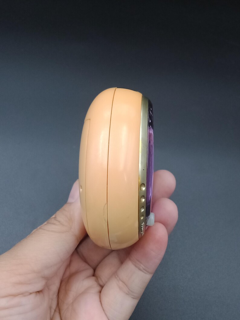 May include: A peach-colored, oval-shaped electronic device with a gold-colored trim. The device has a small, white button and a purple screen. The device is held in a hand against a dark background.