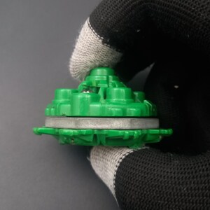 May include: A green plastic toy component with a circular design. The component has a layered structure with a silver-colored ring in the middle. The top section has multiple raised, rounded features. The object is held by a gloved hand against a gray background.
