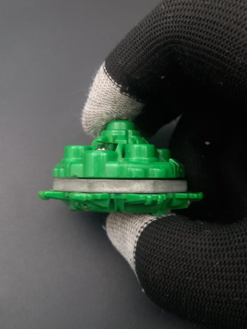 May include: A green plastic toy component with a circular design. The component has a layered structure with a silver-colored ring in the middle. The top section has multiple raised, rounded features. The object is held by a gloved hand against a gray background.