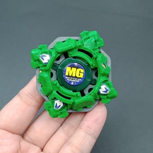 May include: A green and gray toy with a circular design. The toy has green protrusions and a central green circle with the letters "MG" and Japanese text. The toy is held in a hand.