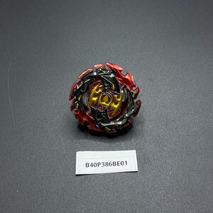 K&ouml;nnte beinhalten: Ein rotes, schwarzes und goldenes Beyblade mit einem scharfen, kantigen Design. Das Beyblade liegt auf einer grauen Oberfl&auml;che mit einem wei&szlig;en Etikett, auf dem "B40P386BE01" steht.