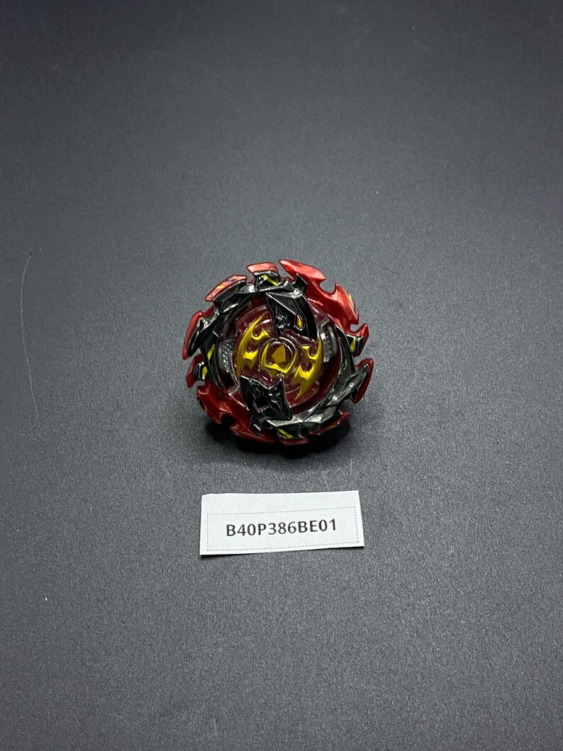 K&ouml;nnte beinhalten: Ein rotes, schwarzes und goldenes Beyblade mit einem scharfen, kantigen Design. Das Beyblade liegt auf einer grauen Oberfl&auml;che mit einem wei&szlig;en Etikett, auf dem "B40P386BE01" steht.