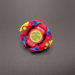 以下が含まれることがあります： 赤、青、黄色のプラスチック製の玩具のトップ。透明なプラスチックの中央に、赤、黄、緑のデザインが施されています。トップはベイブレードです。