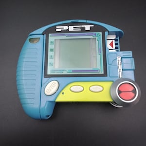 Puede incluir: Un dispositivo electrónico portátil azul y amarillo con la palabra "PET" en la parte superior. Cuenta con una pantalla, botones y un dial circular con detalles en rojo. El dispositivo tiene un diseño retro.