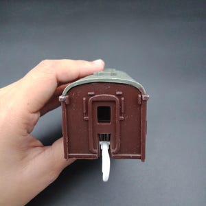 Può includere: Un vagone di treno giocattolo marrone con tetto grigio e connettore bianco. Il vagone ha una forma rettangolare con una piccola apertura al centro. Il vagone del treno giocattolo è una replica in miniatura.