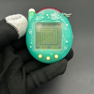 May include: A teal handheld digital pet game with a small screen displaying a pixelated pattern. The device has three yellow buttons, a small antenna, and a red accent. The game is held by a gloved hand.