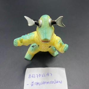May include: A small, green and yellow toy figure with a creature-like design. The figure has large eyes, horns, and outstretched arms. The figure is on a dark surface. A handwritten note is in the image.