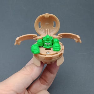 May include: A tan and green toy figure with articulated arms and a spherical body. The figure is open, revealing a green head and arms. The top shell is open, displaying the weight of 380g. The toy is held against a gray background.