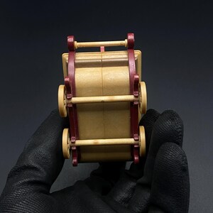 May include: A small, wooden toy wagon with a tan body and burgundy accents. The wagon has four wheels and a handle. The toy is held by a gloved hand against a black background.