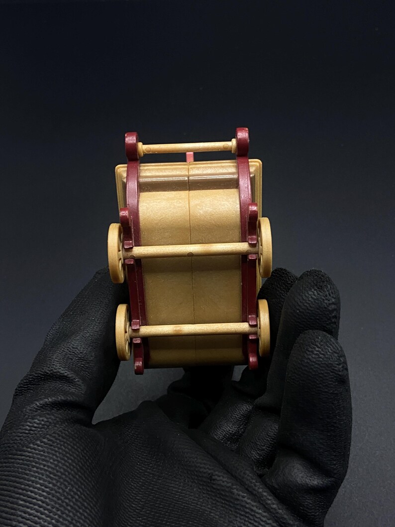 May include: A small, wooden toy wagon with a tan body and burgundy accents. The wagon has four wheels and a handle. The toy is held by a gloved hand against a black background.
