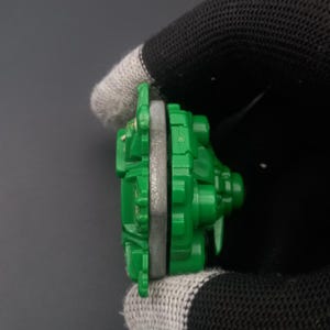 May include: A green and gray toy component, possibly part of a larger set. The toy has a circular shape with a gray ring in the middle and green plastic parts. The object is held by a gloved hand against a dark background.