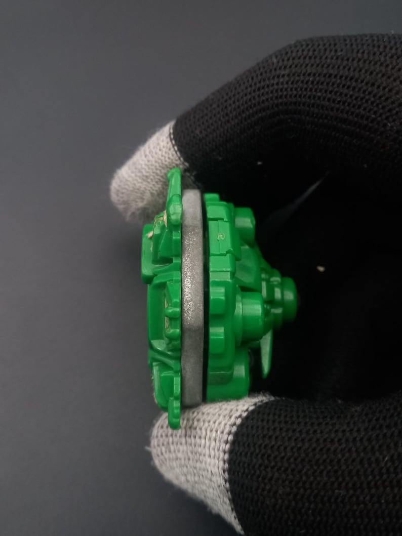 May include: A green and gray toy component, possibly part of a larger set. The toy has a circular shape with a gray ring in the middle and green plastic parts. The object is held by a gloved hand against a dark background.