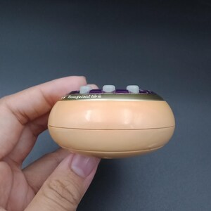 May include: A small, round, peach-colored device with three white buttons and gold trim. The top section has the text "Hungry Tide 2" in gold lettering. The device is held in a hand against a dark gray background.