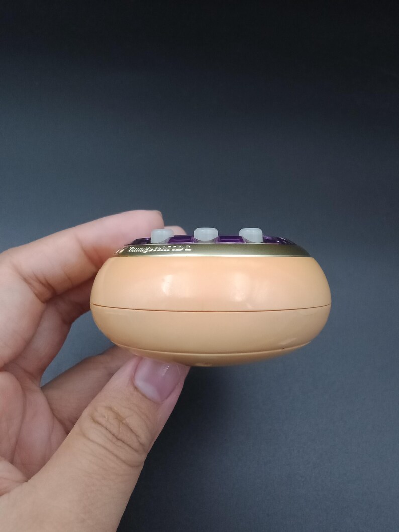 May include: A small, round, peach-colored device with three white buttons and gold trim. The top section has the text "Hungry Tide 2" in gold lettering. The device is held in a hand against a dark gray background.