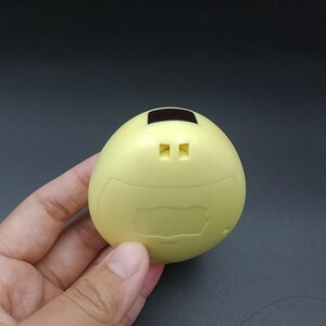 May include: A pale yellow, egg-shaped electronic device with a small rectangular screen and two square buttons. The device has a curved top and a slightly textured surface. The device is held in a hand against a dark background.