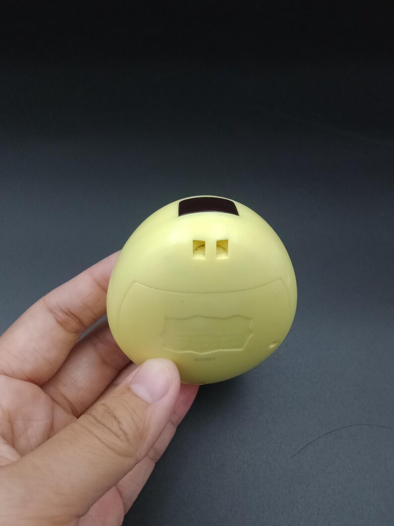 May include: A pale yellow, egg-shaped electronic device with a small rectangular screen and two square buttons. The device has a curved top and a slightly textured surface. The device is held in a hand against a dark background.
