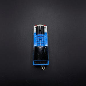 May include: A blue and black toy train engine with a black smokestack and a white face. The engine has a silver hook on the front.