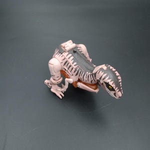 May include: A tan and black toy dinosaur, possibly a Velociraptor, is positioned on a dark gray surface. The toy has a detailed design with visible claws, a textured back, and yellow eyes. The dinosaur is in a crouching pose.