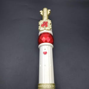 Puede incluir: Una varita decorativa con una parte superior y detalles dorados, una gema roja y una columna blanca con un corazón. La varita tiene una base dorada y está sobre un fondo oscuro. La varita mide aproximadamente 25 cm de largo.