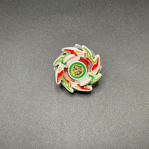 Può includere: Una trottola bianca, rossa e verde con un emblema centrale raffigurante un drago. Il giocattolo ha un design circolare con più lame ed è posto su uno sfondo scuro.