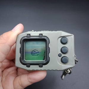 May include: A handheld digital pet game in a gray case with a small screen displaying pixelated graphics. It has three black buttons and a keychain attachment. The screen shows a pixelated landscape with a creature.