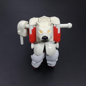 May include: A white and red polar bear transformer toy with yellow eyes and a black nose. The toy has articulated limbs and a detailed bear head. The toy is on a black background.