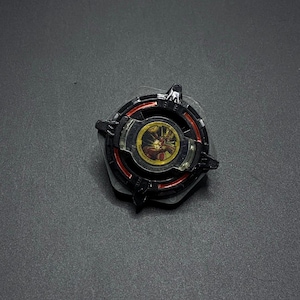 K&ouml;nnte beinhalten: Schwarzes und rotes Kreisel mit einer goldenen und roten Mitte. Die Mitte hat ein Design einer Kreatur mit einem roten und gelben Gesicht.