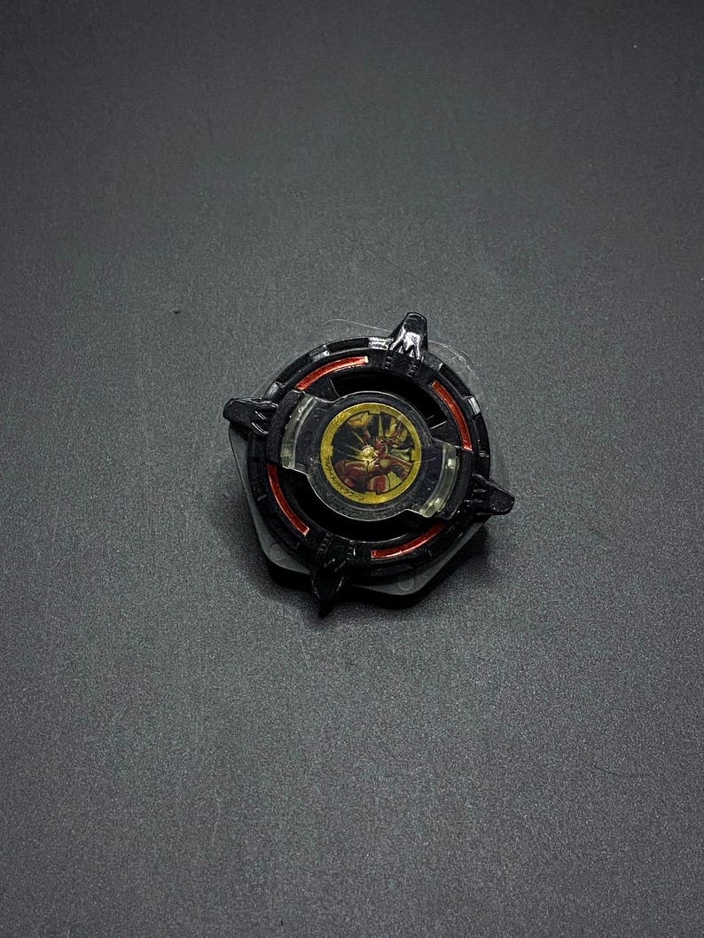 K&ouml;nnte beinhalten: Schwarzes und rotes Kreisel mit einer goldenen und roten Mitte. Die Mitte hat ein Design einer Kreatur mit einem roten und gelben Gesicht.