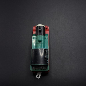 May include: A toy train engine in red, green, and black. The engine has a black smokestack, green body, and red sides. A white hook is visible at the front. The toy is designed for children.