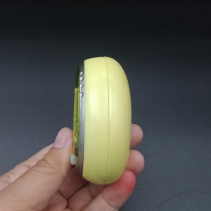 May include: A small, yellow, oval-shaped electronic device with a silver trim and a small white button. The device is held in a hand against a dark background. The device appears to be a timer or a small electronic gadget.