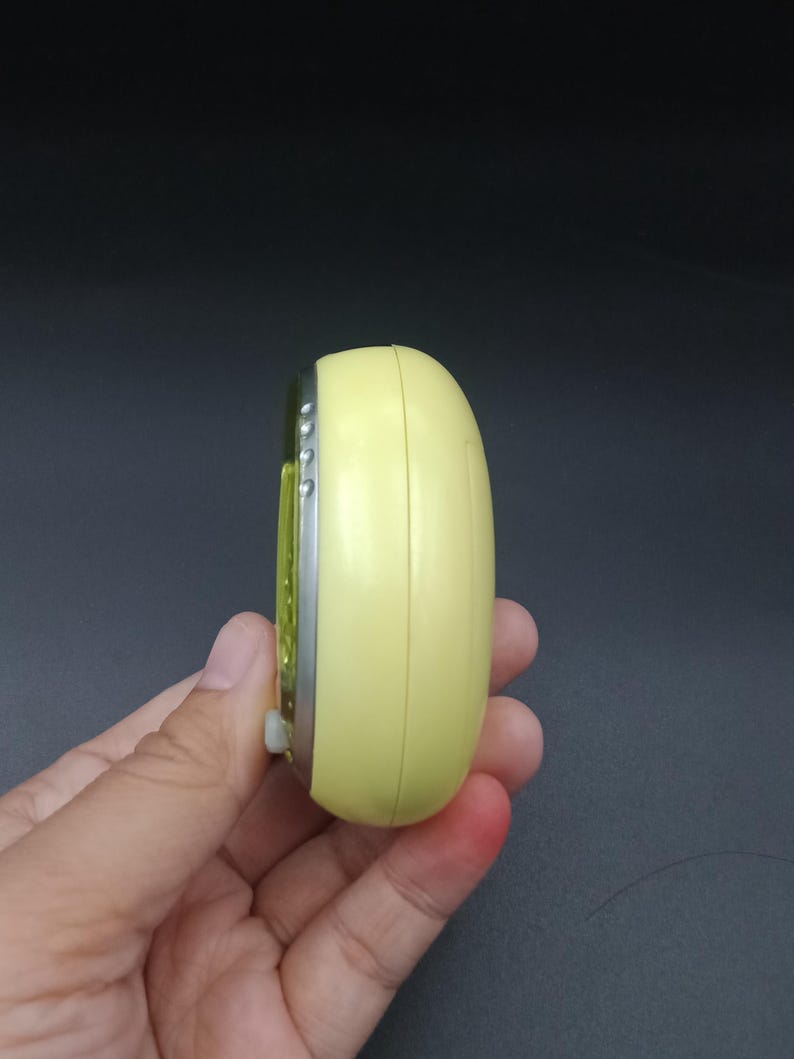 May include: A small, yellow, oval-shaped electronic device with a silver trim and a small white button. The device is held in a hand against a dark background. The device appears to be a timer or a small electronic gadget.