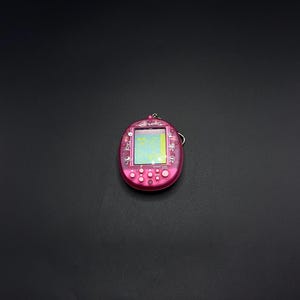 以下が含まれることがあります： ピンク色の携帯型デジタルペットゲーム。小さな画面と複数のボタンがあります。キーホルダーが付いています。画面にはピクセル化されたグラフィックが表示されます。ケースは鮮やかなピンク色で、画面にはカラフルなピクセル化された画像が表示されます。