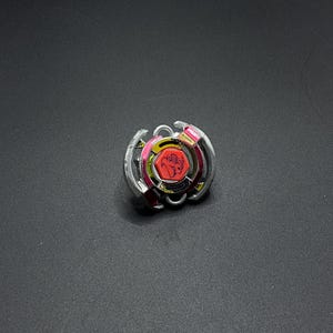 Może przedstawiać: Srebrny i czerwony Beyblade z żółtym i czarnym centrum. Beyblade ma czerwony i srebrny wzór na zewnętrznym pierścieniu.