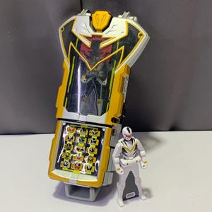 May include: A yellow and silver electronic toy morpher featuring a central display with a small figure inside and a keypad with multiple character icons, accompanied by a small white and yellow action figure with a red visor and black accents.