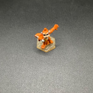 May include: A small, orange monkey figurine with sunglasses and a determined pose. The monkey is standing on a hexagonal base. The figurine has a detailed design and is made of a solid material.