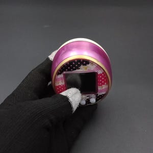 Puede incluir: Un juego electr&oacute;nico port&aacute;til rosa y blanco con una peque&ntilde;a pantalla y dos botones. El dispositivo tiene un borde decorativo con lunares y motivos florales. El dispositivo est&aacute; sostenido por una mano enguantada.